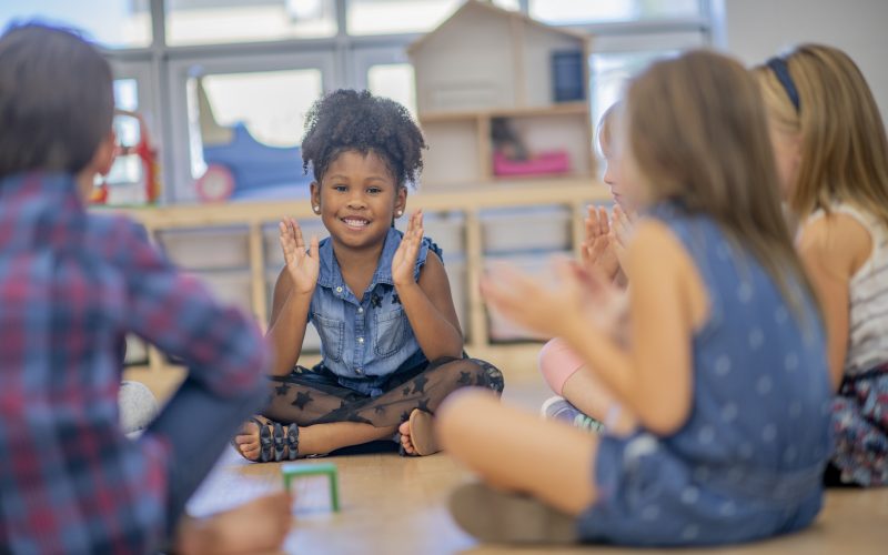 An adorable little girl of african descent sits cross-legged in a circle with her friends. She is clapping and smiling with her classmates.
