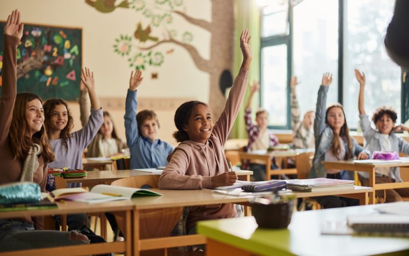 Large group of happy elementary students know the answer on a class in the classroom. Focus is on black girl.
